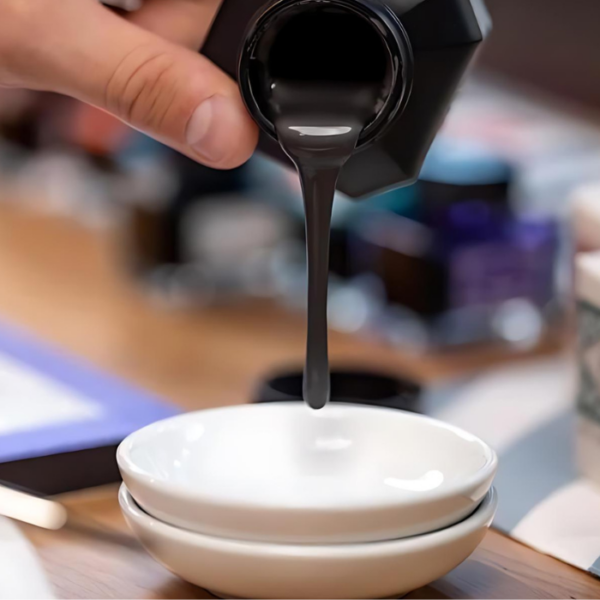 A close up of a persons hand coming in from the left hand side of the frame, pouring some of the Kuretake ZIG Fluid Graphite into a white porcelain bowl. The background is blurred.