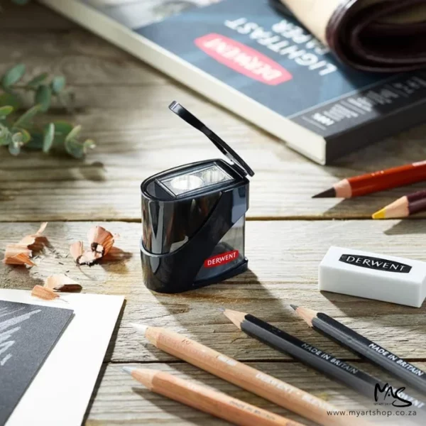 A promotional image for the Derwent Twin Hole Pencil Sharpener. The sharpener is shown in the center of the frame with the black plastic lid open. The sharpener is black and has a red Derwent logo printed on the bottom. It is surrounded by sketch pads and pencils and an eraser. There are some pencil sharpening's next to the sharpener.