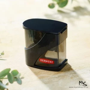 A promotional image of a Derwent Twin Hole Pencil Sharpener. The sharpener is black and has a red logo printed on the bottom. It has a cap over the holes. It is shown at a slight angle in the center of the frame on a wooden surface with some greenery around it.