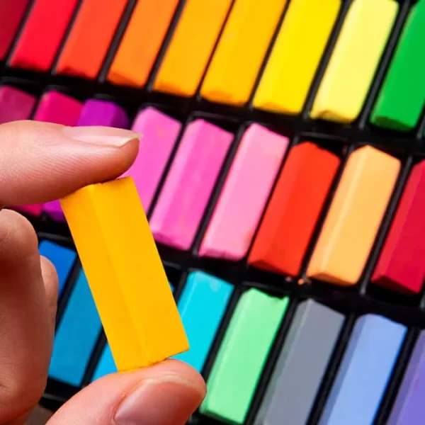 A persons hand is seen coming into the frame, from the bottom left hand side of the frame. They are holding a short, yellow, rectangular Mungyo Sift Pastel in between their fingers. In the background is a set of pastels that form a rainbow and full the frame.