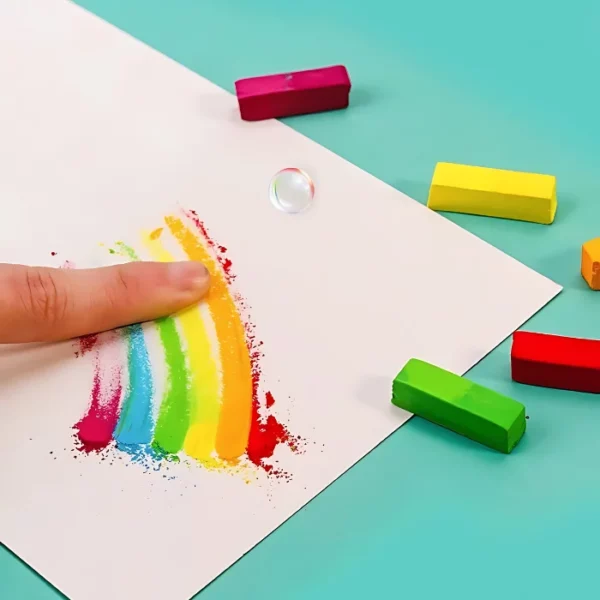 A persons hand is seen coming into the frame in the bottom left hand corner. They are using their finger to smudge a pastel drawing of a rainbow that was made on white paper. There are four Mungyo Soft Pastels laying around the paper. They are red, yellow, pink and green. The pastel sticks are short and rectangular. On a blue background.