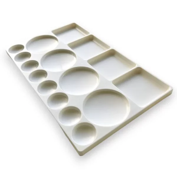 A Prime Art Plastic Palette 16 Well Rectangular is shown, diagonally in the center of the frame. The palette is made of a white plastic and has 16 wells to hold colour. Some of the wells are circular and some are rectangular. The image is center of the frame and on a white background.