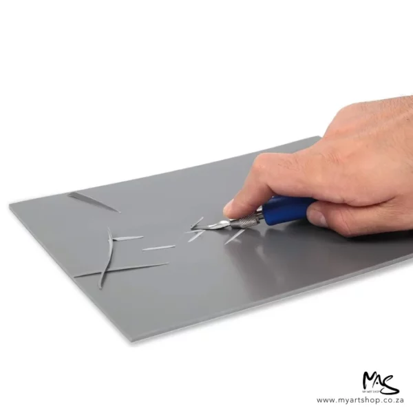 A close up of a persons hand coming in from the right hand side of the frame, carving a piece of lino using a blue handled scraping tool. The lino is grey and slightly angled in the frame. The image is on a white background.