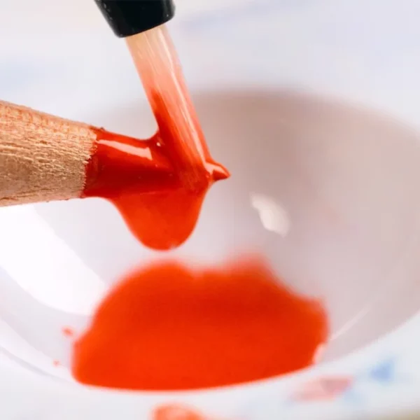 A close up of a single pencil from the Set of 12 Derwent Inktense Pencils. The pencil lead is orange and you can see a waterbrush being dipped onto the lead to gather colour from the lead. There is a pool of orange water under the pencil tip. The image is cut off by the frame.