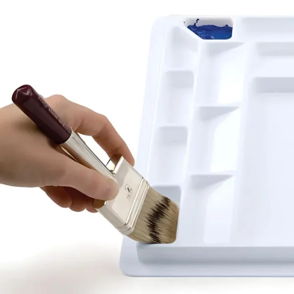 A close up of a persons hand dipping a paint brush into a Sterling Edwards Big Brush Palette. The wells are large enough for big brushes. The palette is made of white plastic and has 14 different wells. On a white background.