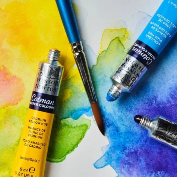 A close up image of three tubes of Winsor and Newton Cotman Watercolour Paints. The paints have their caps off and they are laying on a piece of painted paper. The paper has been painted with yellow and blue washes. There is a Cotman Watercolour brush also laying on the painted surface. The image is cut off by the frame.