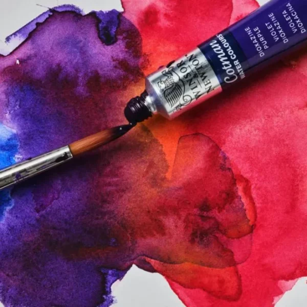 A close up image of a tube of Winsor and Newton Cotman Watercolour Paint shown laying on a piece of painted paper. The lid of the tube is off and you can see the paint coming out of the tube. The paint is purple and is dropping onto a paint brush.