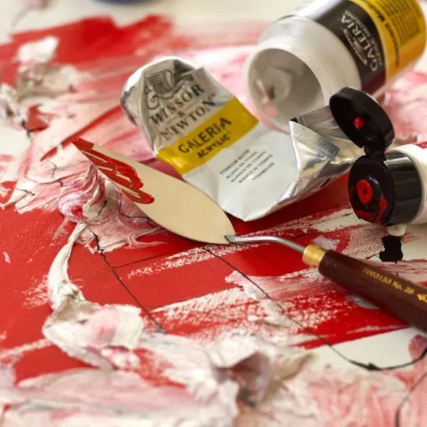 A close up image of some tubes of Winsor and Newton Galeria Acrylic Paint laying on top of a painted canvas. The canvas has red and white paint randomly applied to it and has a lot of texture. There is also a palette knife laying next to the tubes, indicating the paint was applied using the knife. The image is cut off by the frame.