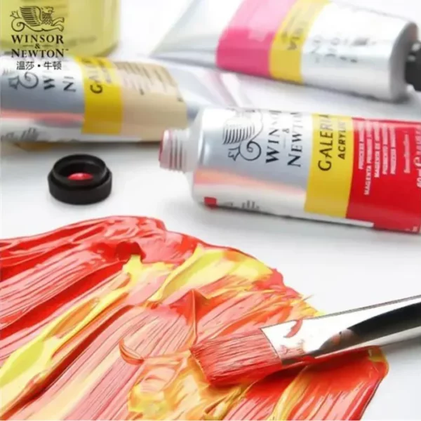 A close up image of some red and yellow Winsor and Newton Galeria Acrylic Paint being applied to a surface. There is a paint brush leaning on top of the paint that has been applied. There are three tubes of Winsor and Newton Galeria Acrylic Paint laying in the background that are slightly blurry. The image is cut off by the frame.