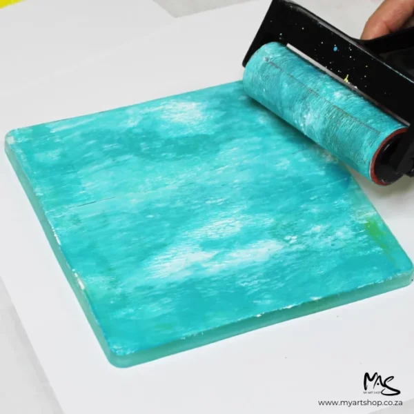 A 6" x 6" Gelli Arts Printing Plate is seen in the center of the frame with a persons hands coming into the frame. They are brayering some aquamarine paint across the printing plate.