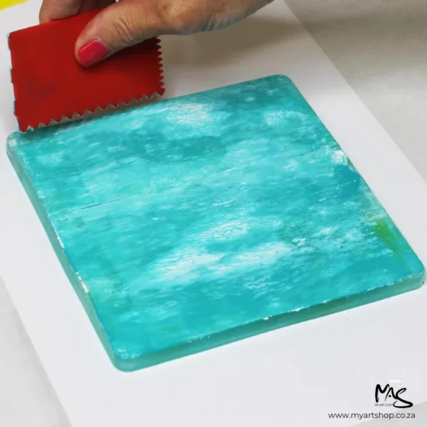 A 6" x 6" Gelli Arts Printing Plate can be seen in the center of the frame. A persons hands are coming into the frame and using a plastic object to run through the paint to create different pattern lines in the paint.