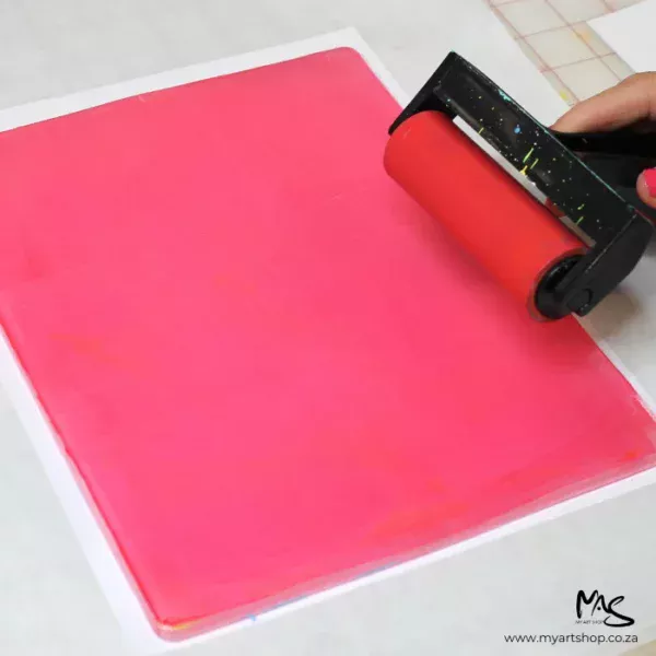 A 8" x 10" Gelli Arts Printing Plate is seen in the center of the frame with a persons hands coming into the frame. They are brayering some pink paint across the printing plate.