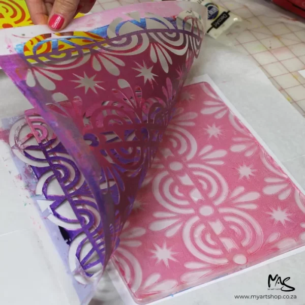 A persons hand is seen lifting the stencil off of the 8" x 10" Gelli Arts Printing Plate which reveals a design the same as the stencil in the pink paint