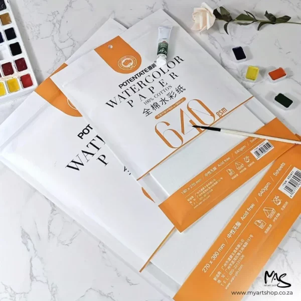 There are two Double Sided Potentate Watercolour Paper Packs shown laying one on top of the other, diagonally across the center of the frame. The image is cut off by the frame. The bigger pack is below the smaller pack. The packs are white with an orange colour band around the base of each packet and text on the packets. There is a paintbrush lying on top of the packs and various watercolour pans scattered around the packs.