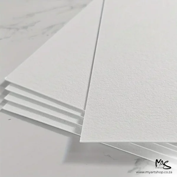 There is a staggered pile of Double Sided Potentate Watercolour Papers shown coming in from the top of the frame, so you can see the corners of each sheet. There are 5 sheets, staggered on top of each other on a marble surface. The paper is thick and white.