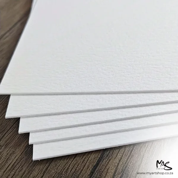 There is a staggered pile of Double Sided Potentate Watercolour Papers shown coming in from the top of the frame, so you can see the corners of each sheet. There are 5 sheets, staggered on top of each other on a wooden surface. The paper is thick and white.