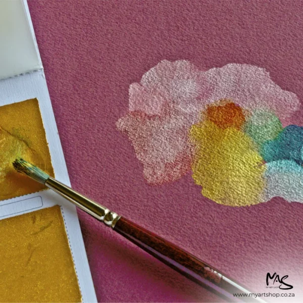 A close up of a paintbrush that is applying some gold paint to a sheet Fabriano Cromia Paper. The image is cut off by the frame. The paper is pink.