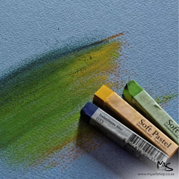 A close up of 3chalk pastels in green, yellow and blue, applying some colour to a sheet of Fabriano Cromia Paper. The image is cut off by the frame. The paper is blue.