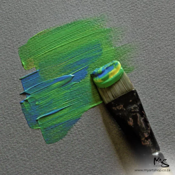A close up of a paintbrush applying some green and blue paint to a sheet of Fabriano Cromia Paper. The image is cut off by the frame. The paper is dark grey.