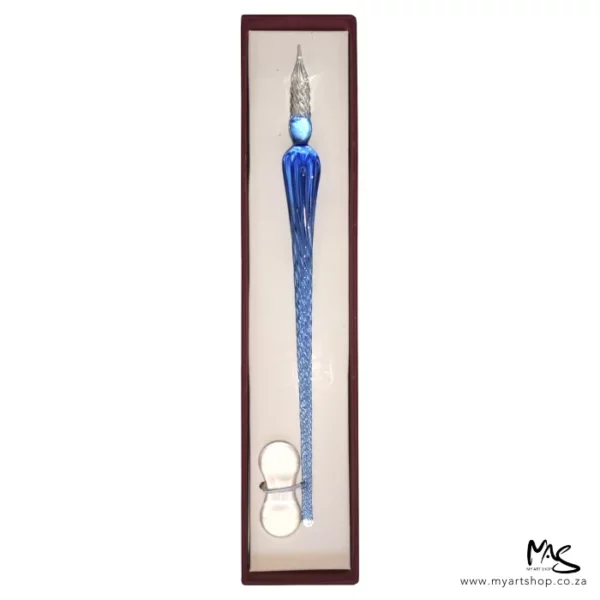 There is a single Glass Dipping Pen Ice Blue in a box, shown vertically, in the center of the frame. The box is brown, and there is a beige cushioning in the bottom of the box and the pen is sat on the cushioning. There is a clear glass pen rest, placed at the bottom of the pen. The image is on a white background.