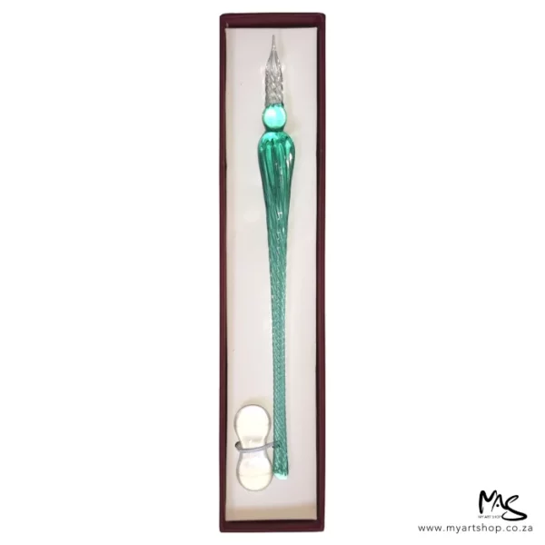 There is a single Glass Dipping Pen Ice Green in a box, shown vertically, in the center of the frame. The box is brown, and there is a beige cushioning in the bottom of the box and the pen is sat on the cushioning. There is a clear glass pen rest, placed at the bottom of the pen. The image is on a white background.
