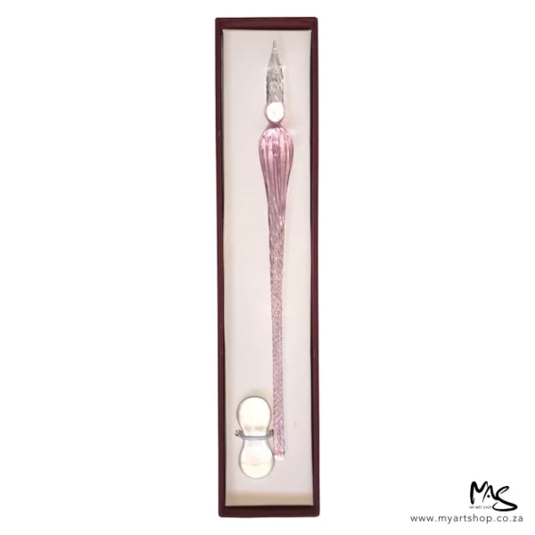 There is a single Glass Dipping Pen Pink in a box, shown vertically, in the center of the frame. The box is brown, and there is a beige cushioning in the bottom of the box and the pen is sat on the cushioning. There is a clear glass pen rest, placed at the bottom of the pen. The image is on a white background.