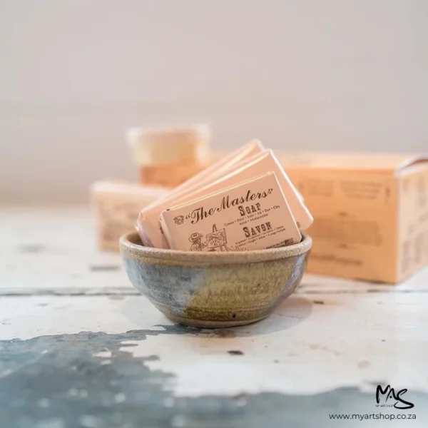 There is a ceramic bowl in the center of the frame. There are a few bars of Masters Hand Soap inside the bowl. The soap is in its beige paper packaging and there is brown text on the wrapper. There are more bars in the background that are blurred. The bowl is sitting on a weathered stone surface. The image is cut off by the frame.