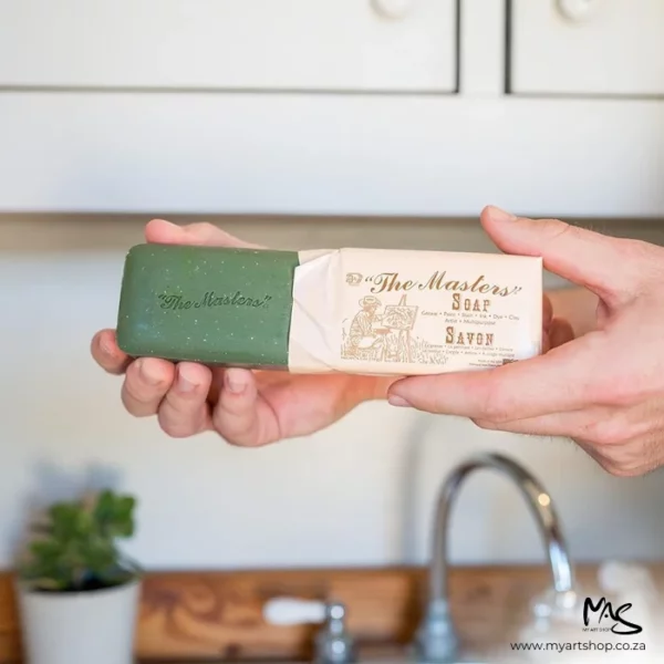 A persons hands are seen coming in from the right hand side of the frame, they are removing the paper wrapper from a bar of Masters Hand Soap. The packaging is beige with brown text and the bar is green. There is a kitchen sink in the background that is blurred.
