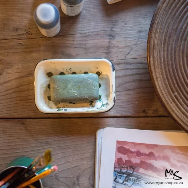 There is a bar of Masters Hand Soap sitting in a little metal bowl in the center of the frame. The soap has been used. There are some brushes in a bowl nearby and some watercolour paintings in the bottom right hand corner of the frame that are cut off by the frame. The image is a birds eye view and there is an old wooden table under the products.