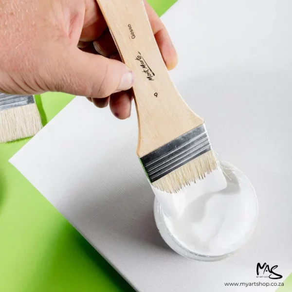 A single brush from the Mont Marte Discovery Gesso Brush Set is seen in a persons hand, which is coming in from the left hand side of the frame. They are applying white gesso to a white canvas. There is a green background and the image is cut off by the frame. It is a birds eye view.