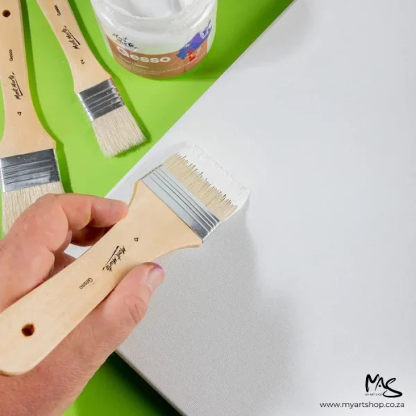 The 3 loose brushes from the Mont Marte Discovery Gesso Brush Set are seen in this frame. Two brushes are laying on the surface and a persons hand is coming in from the bottom left hand corner of the frame holding a brush and applying white gesso to a canvas. You can see part of the open jar of gesso in the top of the frame. The image is cut off by the frame. It is a birds eye view and there is a bright green background.
