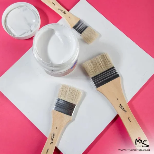 The 3 brushes from the Mont Marte Discovery Gesso Brush Set are shown laying on top of a piece of paper with an open jar of gesso. The image has a bright pink background. It is a birds eye view. The image is cut off by the frame.