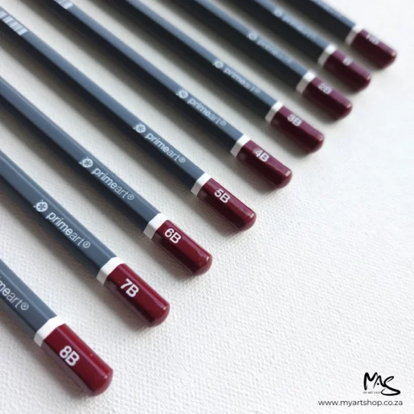 Prime Art Graphite Pencil Set. 9 pencils are laying diagonally across the center of the frame. The pencils have a maroon tip with the grade printed in white and a dark grey barrel. The maroon ends are facing the right hand side of the frame and the lead tips are facing towards the top left hand corner. They are on a light grey background with a canvas texture.
