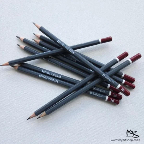 Prime Art Graphite Pencil Set. A pile of pencils is shown in the center of the frame. The pencils are laying randomly on top of each other at different angles. All the ends of the pencils are facing towards the right hand side of the frame and the lead tips are facing towards the left hand side of the frame. The pencils have a dark grey barrel and a maroon tip with the grade printed on the tip. They are center of the frame and on a light grey background.