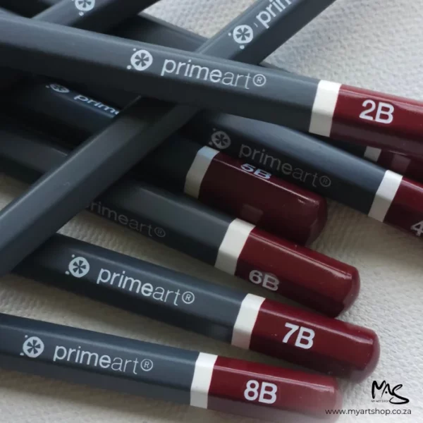 Prime Art Graphite Pencil Set. A close up of a pile of pencils laying randomly on top of each other. The pencil tips are showing with part of the barrel. The tips are maroon in colour and have the grade of the pencil printed in white. The barrel of the pencils are a dark grey and the prime art brand is printed in white.