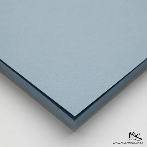 A close up of the corner of a pile of papers from the Sea Fabriano Toned Paper Pad. The corner of the papers is pointing towards the center of the frame. The paper is blue and on a light grey background.