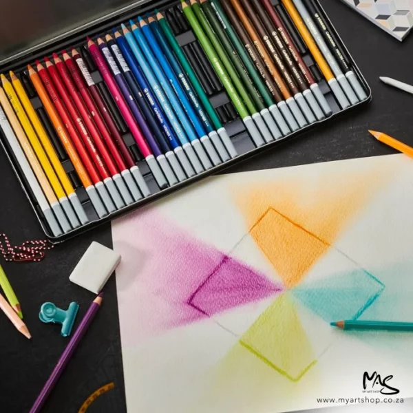 A promotional image of a Stabilo CarbOthello Chalk Pastel Pencil Set. The set is shown diagonally across the back of the frame and there is a sheet of paper in the front of the frame that has been drawn on using the pencils. There are different shades shown in a square block. There are various pencils laying around the set and the paper.