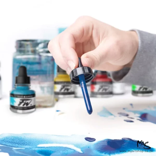 A promotional image for Daler Rowney FW Acrylic Inks. A close up of a persons hand holding the eyedropper from a blue ink and dropping ink onto a piece of paper. There are other bottles of ink in the background that are blurred. The image is cut off by the frame.