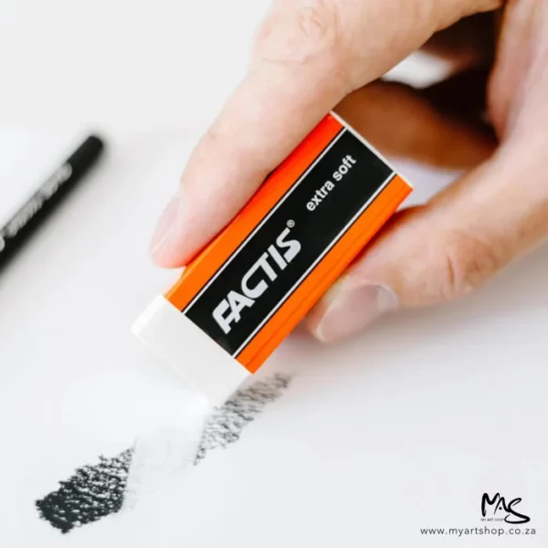 A close up of a persons hand holding a Factis Extra Soft Eraser. The eraser is white plastic and is in a black and red printed sleeve with the Factis logo printed on it. They are erasing a pencil drawing on white paper. The image is cut off by the frame.
