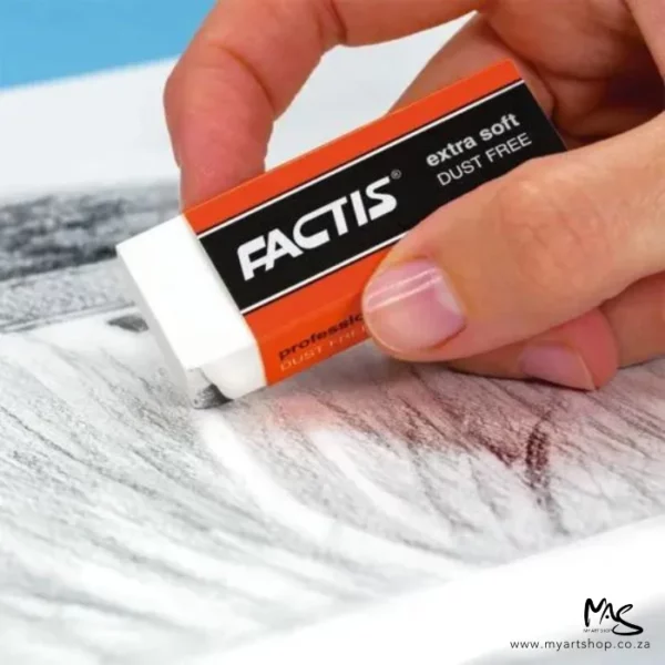 A close up of a persons hand holding a Factis Extra Soft Eraser. The eraser is white plastic and is in a black and red printed sleeve with the Factis logo printed on it. They are erasing a pencil drawing on white paper and the background is blue. The image is cut off by the frame.