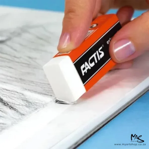 A close up of a persons hand holding a Factis Extra Soft Eraser. The eraser is white plastic and is in a black and red printed sleeve with the Factis logo printed on it. They are erasing a pencil drawing on white paper and the background is blue. The image is cut off by the frame.