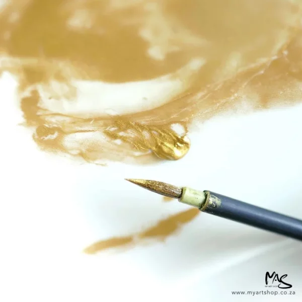 A close up of a paint brush with Gold Kuretake Mica Ink on the bristles. The brush is laying on top of a white piece of paper that has gold ink swirling over it.