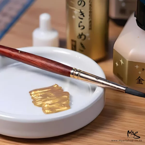 A close up of some Gold Kuretake Mica Paste that has been put in a white ceramic bowl. There is a paint brush leaning across the bowl. Parts of the paste bottle can be seen in the background.