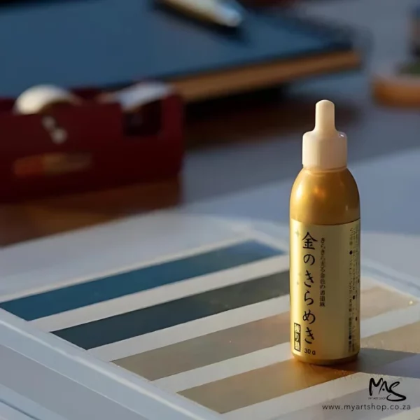 A bottle of Gold Kuretake Mica Paste is seen standing on a colour chart. The bottle is clear plastic and has a clear plastic screw on lid over the nozzle. There is a gold paste inside the bottle and there is a gold label around the body of the bottle with black chinese writing. The background is blurred.