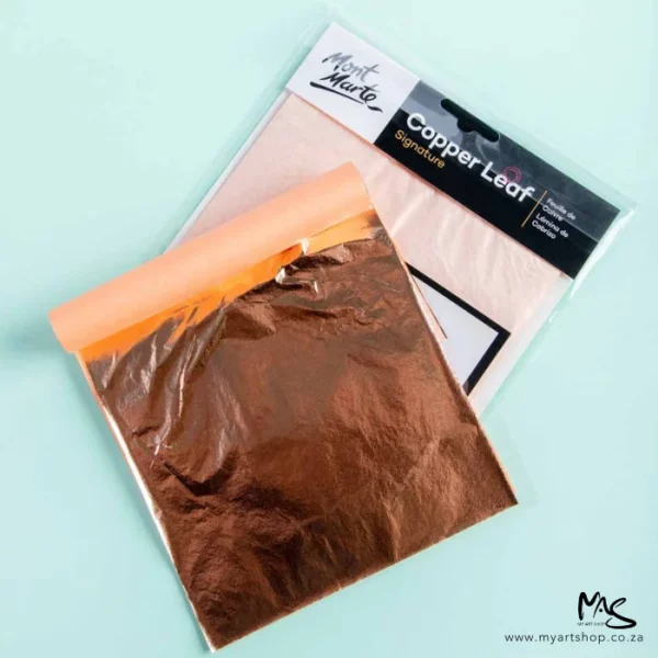 A sealed pack of Mont Marte Copper Leaf and an open pack can be seen laying on top of each other in the center of the frame. The sealed pack is at the back and the open pack is on the top. You can see the copper sheet of the open pack. They are laying on a turquoise background.