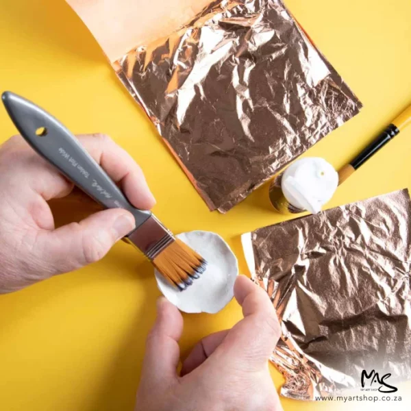 A promotional image for Mont Marte Copper Leaf. The image is taken from a birds eye view. There is a yellow background and a persons hands can be seen holding a brush and applying size to a small white clay pot. There are open pads of copper leaf around their hands.