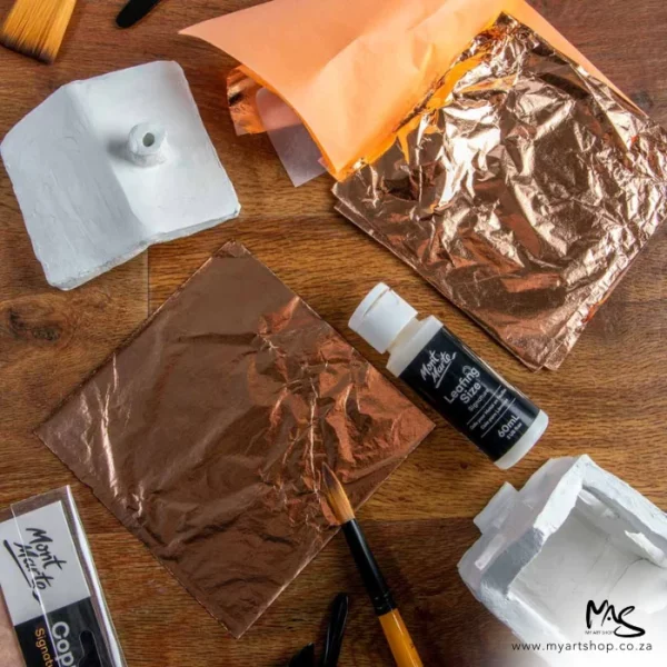 A promotional image for Mont Marte Copper Leaf. It is a birds eye view. There is a wooden background and there are 2 pads of copper leaf laying on the surface with a bottle of leafing size and a paint brush.