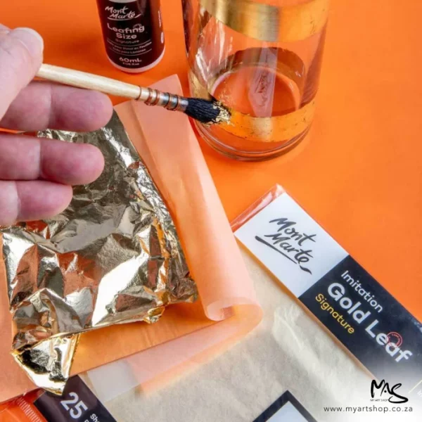A persons hand can be seen coming in from the left hand side of the frame. They are holding a paint brush with gold leaf on the bristles. There is a glass and some pads on the table behind their hand. The image has an orange background.