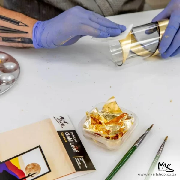 A close up of a persons hand applying Mont Marte Imitation Gold Leaf to a glass. There is a pack of gold leaf on the table and a glass bowl filled with crushed gold leaf and some paint brushes in the frame. The image is cut off by the frame.