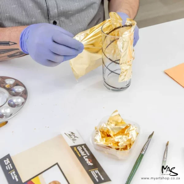 A close up of a persons hand applying Mont Marte Imitation Gold Leaf to a glass. There is a pack of gold leaf on the table and a glass bowl filled with crushed gold leaf and some paint brushes in the frame. The image is cut off by the frame.